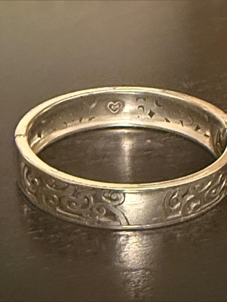 May include: A silver-toned hinged bangle bracelet featuring an embossed swirling pattern on the exterior surface. The interior of the metal band displays engraved celestial motifs including a small heart, crescent moons, and four-pointed stars.