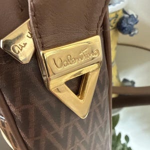 May include: A brown handbag featuring a repeating V-shaped pattern on the fabric. The bag includes a gold-toned triangular metal hardware piece embossed with the word Valentino, and a matching gold-toned zipper pull with partial text.