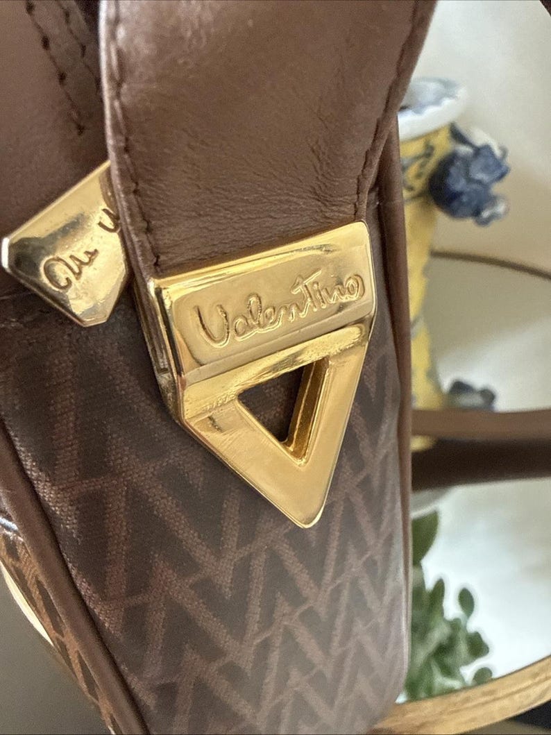 May include: A brown handbag featuring a repeating V-shaped pattern on the fabric. The bag includes a gold-toned triangular metal hardware piece embossed with the word Valentino, and a matching gold-toned zipper pull with partial text.