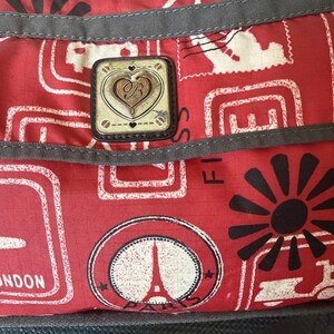May include: Red travel bag with a repeating pattern of white and black shapes, including the Eiffel Tower and the word "London". A decorative emblem with a heart and the letter "B" is centered on the bag.