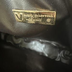 May include: A dark brown interior lining of a bag featuring a rectangular gold-toned metal plaque. The plaque displays the letter V, the text MARIO VALENTINO, the text MADE IN ITALY, and a registered trademark symbol in a clear, embossed font.