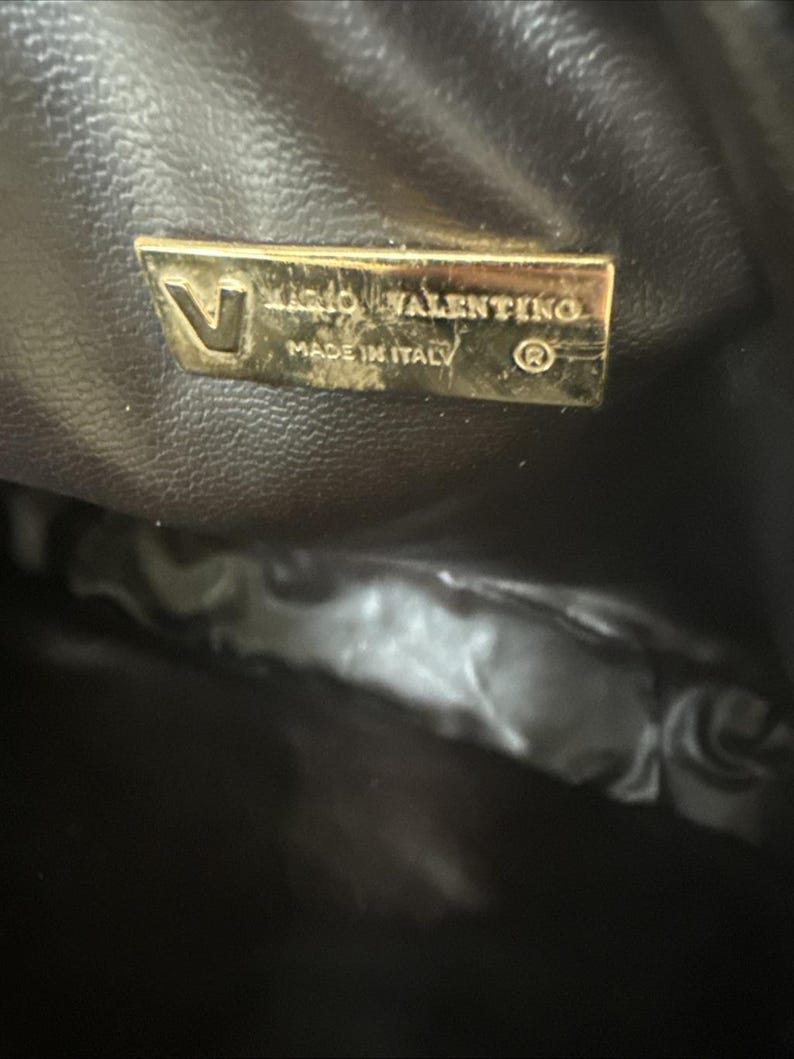May include: A dark brown interior lining of a bag featuring a rectangular gold-toned metal plaque. The plaque displays the letter V, the text MARIO VALENTINO, the text MADE IN ITALY, and a registered trademark symbol in a clear, embossed font.