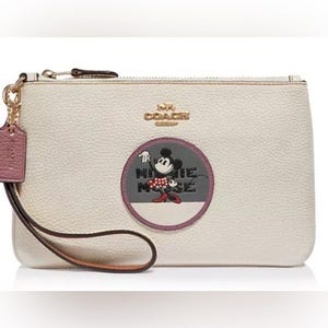 May include: Cream-colored Coach wristlet with a Minnie Mouse graphic. The wristlet features a gold zipper, a wrist strap, and a small tag. The Minnie Mouse graphic is in a circular design with the words "Minnie Mouse".