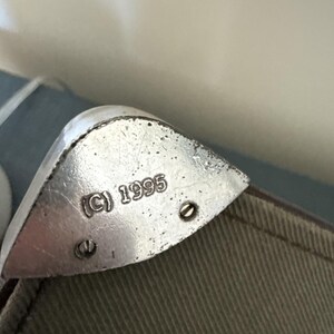 May include: Close-up of a silver metal corner protector with the copyright symbol and the year 1995 stamped on it. The corner protector is attached to a khaki-colored fabric with two small screws.