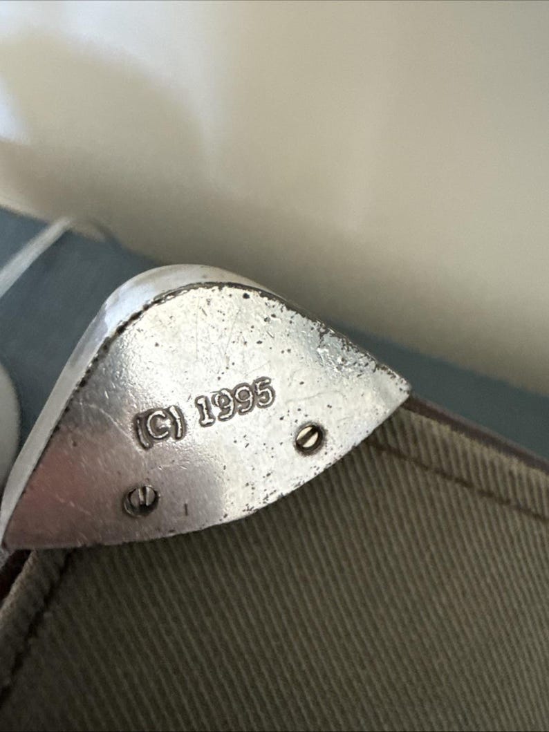 May include: Close-up of a silver metal corner protector with the copyright symbol and the year 1995 stamped on it. The corner protector is attached to a khaki-colored fabric with two small screws.