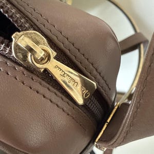 May include: A taupe-colored leather handbag featuring a gold-tone metal zipper pull engraved with the text Mario Valentino. The bag has a smooth, matte finish with visible stitching along the seams and a matching leather strap attached to the side.