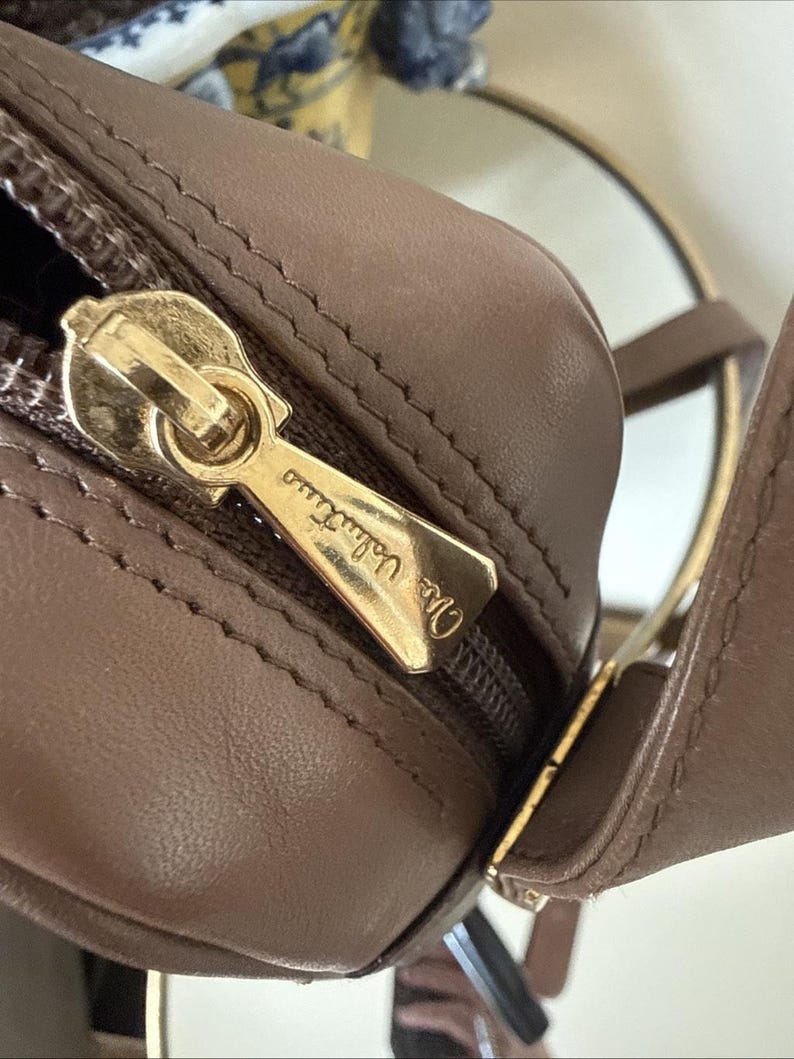 May include: A taupe-colored leather handbag featuring a gold-tone metal zipper pull engraved with the text Mario Valentino. The bag has a smooth, matte finish with visible stitching along the seams and a matching leather strap attached to the side.