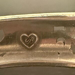 May include: A metallic bracelet featuring a cutout design and a small heart-shaped emblem engraved with the letter B. The piece has a polished finish and a curved band structure, showcasing a distinct hallmark detail on the interior surface.