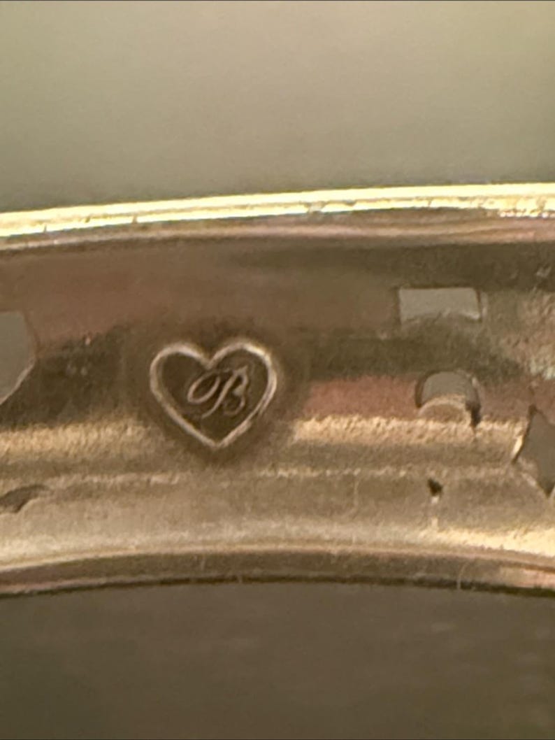 May include: A metallic bracelet featuring a cutout design and a small heart-shaped emblem engraved with the letter B. The piece has a polished finish and a curved band structure, showcasing a distinct hallmark detail on the interior surface.