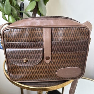 May include: A brown rectangular shoulder bag featuring a repeating V-pattern across the body. The bag includes a front pocket with a gold-toned circular snap closure, a central vertical leather strap with a snap, and a leather patch with text.