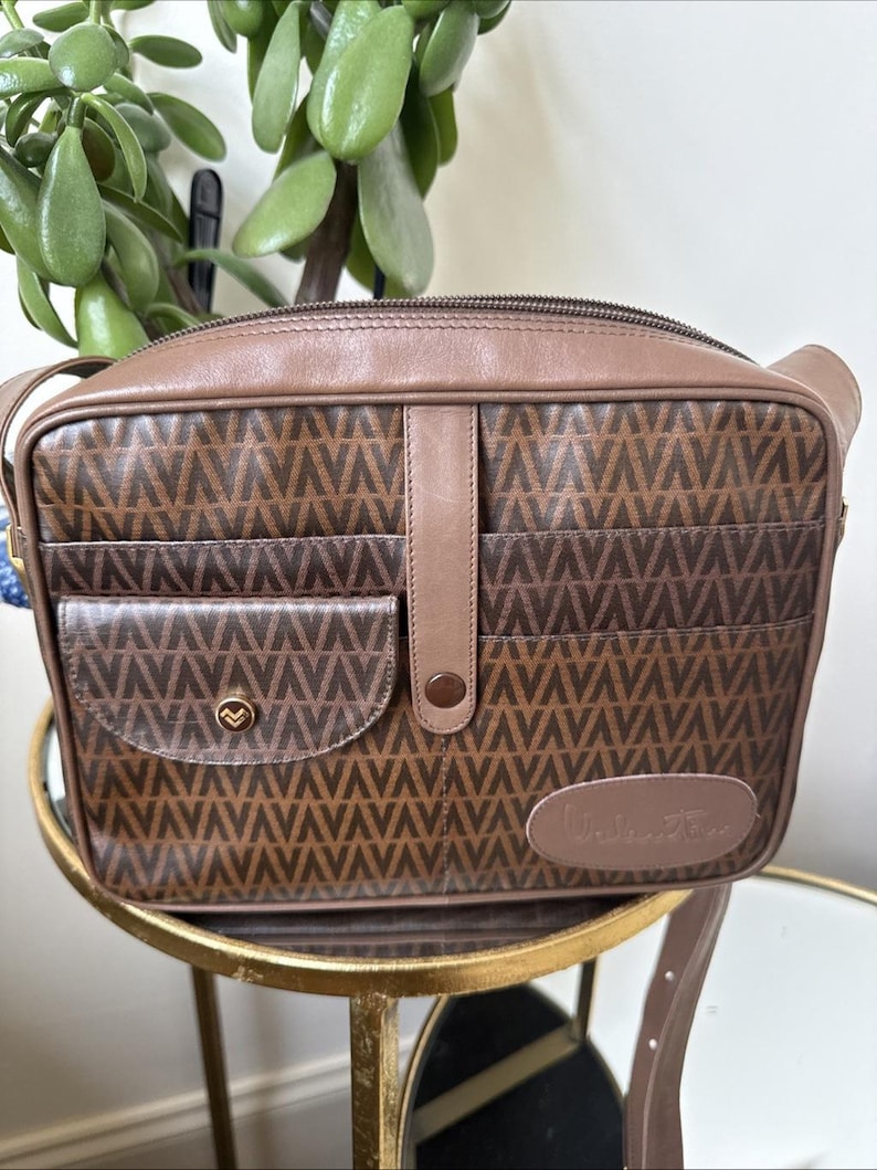 May include: A brown rectangular shoulder bag featuring a repeating V-pattern across the body. The bag includes a front pocket with a gold-toned circular snap closure, a central vertical leather strap with a snap, and a leather patch with text.
