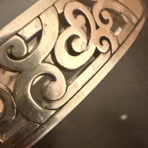 May include: A silver-toned metal cuff bracelet featuring an openwork design with repeating curved, scroll-like patterns. The surface of the metal has a polished finish with visible light reflections along the edges of the intricate cutouts.