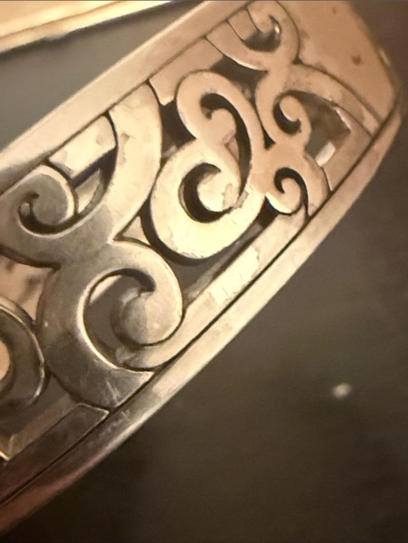 May include: A silver-toned metal cuff bracelet featuring an openwork design with repeating curved, scroll-like patterns. The surface of the metal has a polished finish with visible light reflections along the edges of the intricate cutouts.