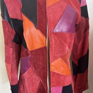 Può includere: Giacca patchwork con una varietà di colori tra cui rosso, nero, arancione e viola. La giacca ha una chiusura con cerniera ed è realizzata in pelle e camoscio. Il design è un mix di forme geometriche.