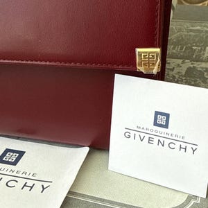 May include: A deep red leather handbag featuring a gold-toned square metal emblem with a geometric logo. Two white cards are present, each displaying the text MAROQUINIERIE GIVENCHY above a square logo. The handbag has a smooth, matte finish.