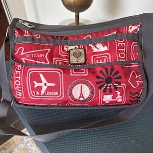 May include: Red crossbody bag with a travel-themed print featuring stamps, an airplane, and the Eiffel Tower. The bag has a gray strap and trim, a zippered top, and a decorative emblem.