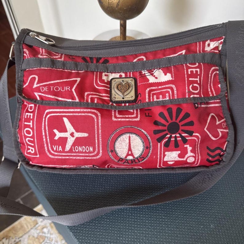 May include: Red crossbody bag with a travel-themed print featuring stamps, an airplane, and the Eiffel Tower. The bag has a gray strap and trim, a zippered top, and a decorative emblem.