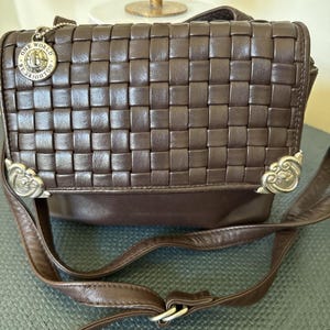 May include: A brown leather crossbody bag with a woven design on the front flap. The bag has a long, adjustable strap and decorative metal accents. A small charm with the words "ONE WORLD BY BRIGHTON" hangs from the top.