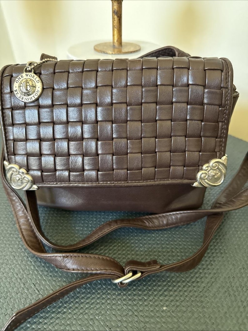 May include: A brown leather crossbody bag with a woven design on the front flap. The bag has a long, adjustable strap and decorative metal accents. A small charm with the words "ONE WORLD BY BRIGHTON" hangs from the top.