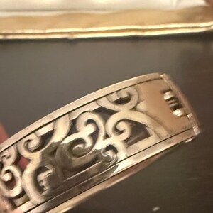 May include: A silver-toned metal hinged bangle bracelet featuring an intricate cut-out scrollwork pattern across the outer surface. The bracelet has a polished finish and a visible hinge mechanism on the side for opening and closing the piece.
