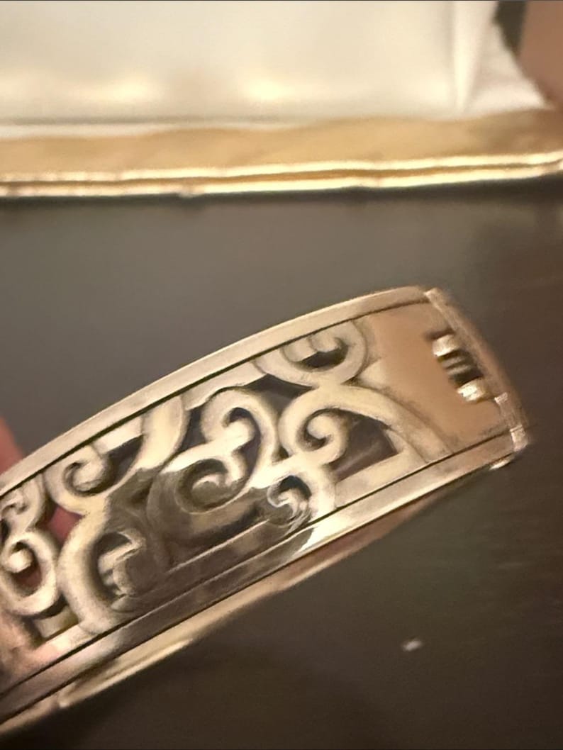 May include: A silver-toned metal hinged bangle bracelet featuring an intricate cut-out scrollwork pattern across the outer surface. The bracelet has a polished finish and a visible hinge mechanism on the side for opening and closing the piece.