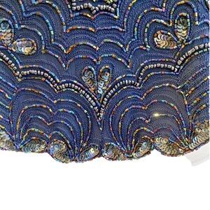Op de afbeelding: Close-up van een marineblauwe stof versierd met iriserende pailletten en kralen. Het ontwerp toont een herhalend patroon van golvende lijnen en cirkelvormige accenten, waardoor een gestructureerd en decoratief oppervlak ontstaat. De pailletten en kralen zorgen voor een glinsterend effect.
