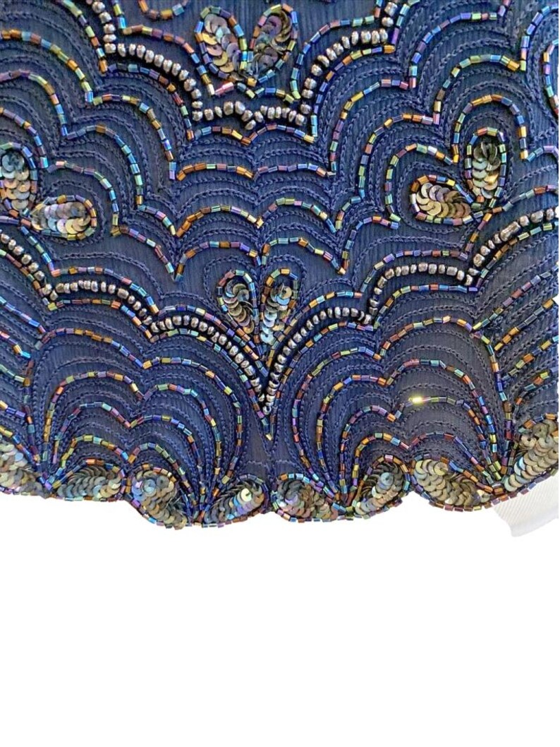 Op de afbeelding: Close-up van een marineblauwe stof versierd met iriserende pailletten en kralen. Het ontwerp toont een herhalend patroon van golvende lijnen en cirkelvormige accenten, waardoor een gestructureerd en decoratief oppervlak ontstaat. De pailletten en kralen zorgen voor een glinsterend effect.