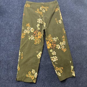 May include: Olive green trousers featuring a repeating floral pattern in shades of yellow, orange, and cream. The trousers have a straight-leg silhouette with a clean waistband and small side slits at the bottom hem of each leg, showcasing a botanical print.