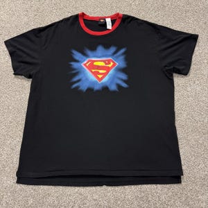May include: A black short-sleeved t-shirt with a red crew neckline. The centre of the chest features a red and yellow Superman logo emblem surrounded by a blue starburst graphic. The shirt includes a small interior tag at the back of the neck.