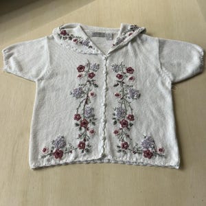 May include: A white short-sleeved knit cardigan featuring a wide collar and a button-down front. The garment is decorated with embroidered floral patterns in shades of pink, purple, and green, extending from the collar down to the hem.