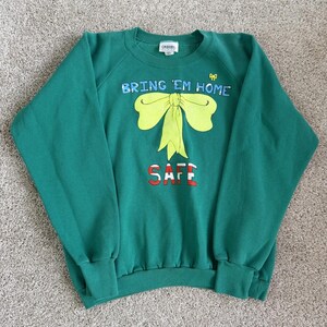 May include: A forest green crewneck sweatshirt featuring a large yellow bow graphic in the centre. The text "BRING 'EM HOME" is printed in blue above the bow, and the word "SAFE" is printed in red below the bow. A small yellow bow icon is visible.