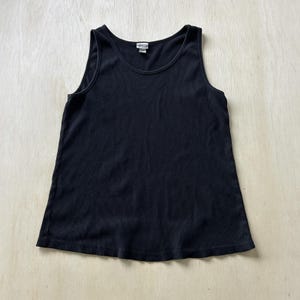 May include: A solid black sleeveless tank top featuring a ribbed texture throughout the fabric, a rounded scoop neckline, and a straight hemline. A small white brand label is visible at the center of the inner neckline collar area.