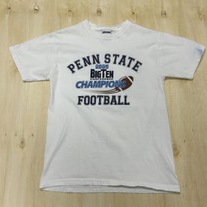 May include: White t-shirt with the text "PENN STATE 2005 BIG TEN CONFERENCE CHAMPIONS FOOTBALL" in blue and brown. The shirt has short sleeves and a crew neck.