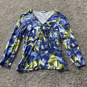 May include: A long-sleeve V-neck top featuring a vibrant floral pattern in shades of blue, green, and white. The garment includes a central twist detail at the chest, creating gathered fabric folds. A small label at the neckline displays the text Cache.