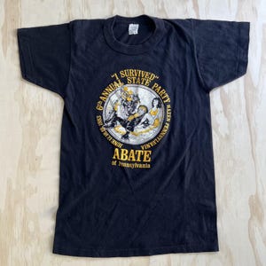 May include: A black short-sleeve t-shirt featuring a circular graphic in yellow and white. The text reads I SURVIVED 6TH ANNUAL STATE PARTY JUNE 17, 18, 19 1983 HAZEN PENNSYLVANIA ABATE of Pennsylvania, surrounding an illustration of two figures.