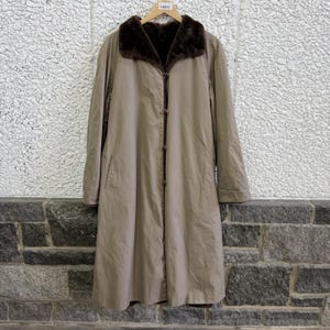 May include: A long, beige coat featuring a dark brown faux fur collar and a front button closure. The coat has long sleeves and a straight silhouette, hanging from a wooden hanger with a small tag labeled 3-8832 at the neckline.