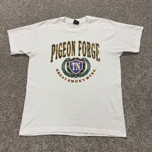 May include: A white short-sleeve t-shirt featuring a graphic on the chest with the text PIGEON FORGE in a patterned font, a central shield emblem containing the letters TN, and the text GREAT SMOKY MTNS. below the shield design.