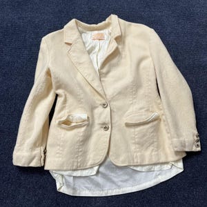 May include: A cream-colored blazer featuring a textured fabric, a notched lapel, and two front buttons. The garment includes two flap pockets, long sleeves with three small buttons at each cuff, and a white satin-like lining that extends below the hem.