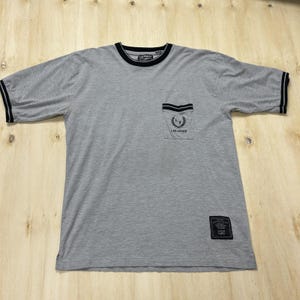 May include: A heather gray short-sleeve t-shirt with black trim on the crew neckline and sleeve cuffs. It features a chest pocket with a black and white striped top edge, a laurel wreath logo, and the text LAS VEGAS. A black rectangular patch is at the hem.