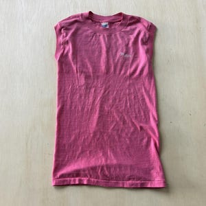 May include: A sleeveless pink t-shirt with a crew neckline. The shirt features the word Maui printed in a small white script font on the upper left chest area. The garment has a straight hemline and a simple, solid-coloured design throughout.