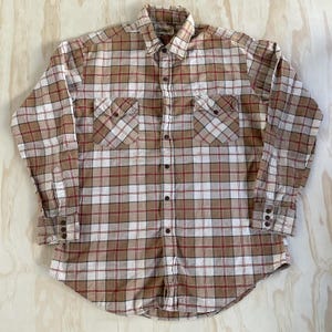 May include: A long-sleeve button-down shirt featuring a plaid pattern in shades of tan, white, and dark red. The shirt includes a pointed collar, two chest pockets with buttoned flaps, and cuffs with three buttons each. The fabric has a visible texture.
