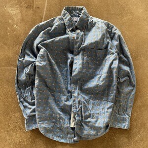 May include: A long-sleeve button-down shirt in a faded blue colour featuring a repeating pattern of small tan and brown figures. The shirt has a standard collar, a single chest pocket on the left side, and a GAP brand label on the inner neck collar.