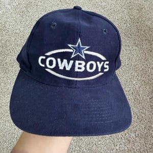 May include: A navy blue baseball cap featuring an embroidered white oval outline surrounding the word COWBOYS in white capital letters, with a blue and white star positioned directly above the text. The cap has a structured crown and brim.