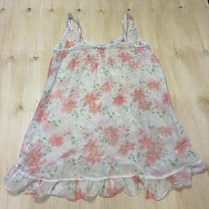 May include: A sleeveless white camisole top featuring a repeating pattern of soft pink flowers and small green leaves. The garment has a scoop neckline, thin shoulder straps, a small button placket at the center chest, and a ruffled hemline.