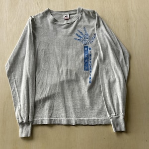 Peut inclure: Un t-shirt à manches longues gris clair avec un motif bleu représentant une main et le texte "BIG AIR WINDSURFING" imprimé verticalement en dessous. Le t-shirt possède un col rond et des poignets côtelés, avec une étiquette de marque visible.