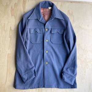 May include: A periwinkle blue button-up jacket featuring a pointed collar, long sleeves with cuffs, and two chest pockets with pointed flaps. The garment has a front button closure with four visible buttons and a textured, woven fabric finish.