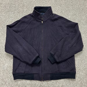 May include: A dark navy blue zip-up jacket featuring a high collar, long sleeves with ribbed cuffs, and a ribbed hem. The jacket is constructed from a textured fabric and includes a central metal zipper closure that extends up to the neck.