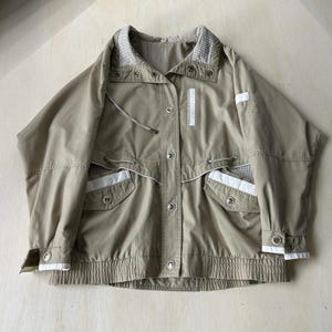 May include: A tan jacket with white accents and a checkered collar. The jacket features a drawstring waist, button closures, and pockets. The cuffs and pockets are trimmed with white. The jacket has a vintage aesthetic.