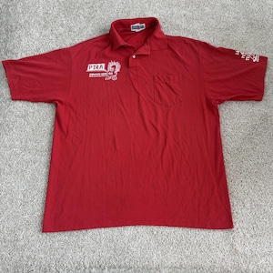 May include: A red polo shirt with a collar, pocket, and short sleeves. The shirt has white text and a graphic on the front, and text on the sleeve. The shirt is made of a soft material.