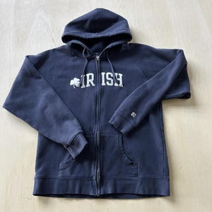 Puede incluir: Sudadera con capucha azul marino con cremallera y bolsillos delanteros. La palabra "IRISH" está impresa en letras blancas en el pecho, con un trébol en el lado izquierdo. Un pequeño logotipo está en la manga derecha.
